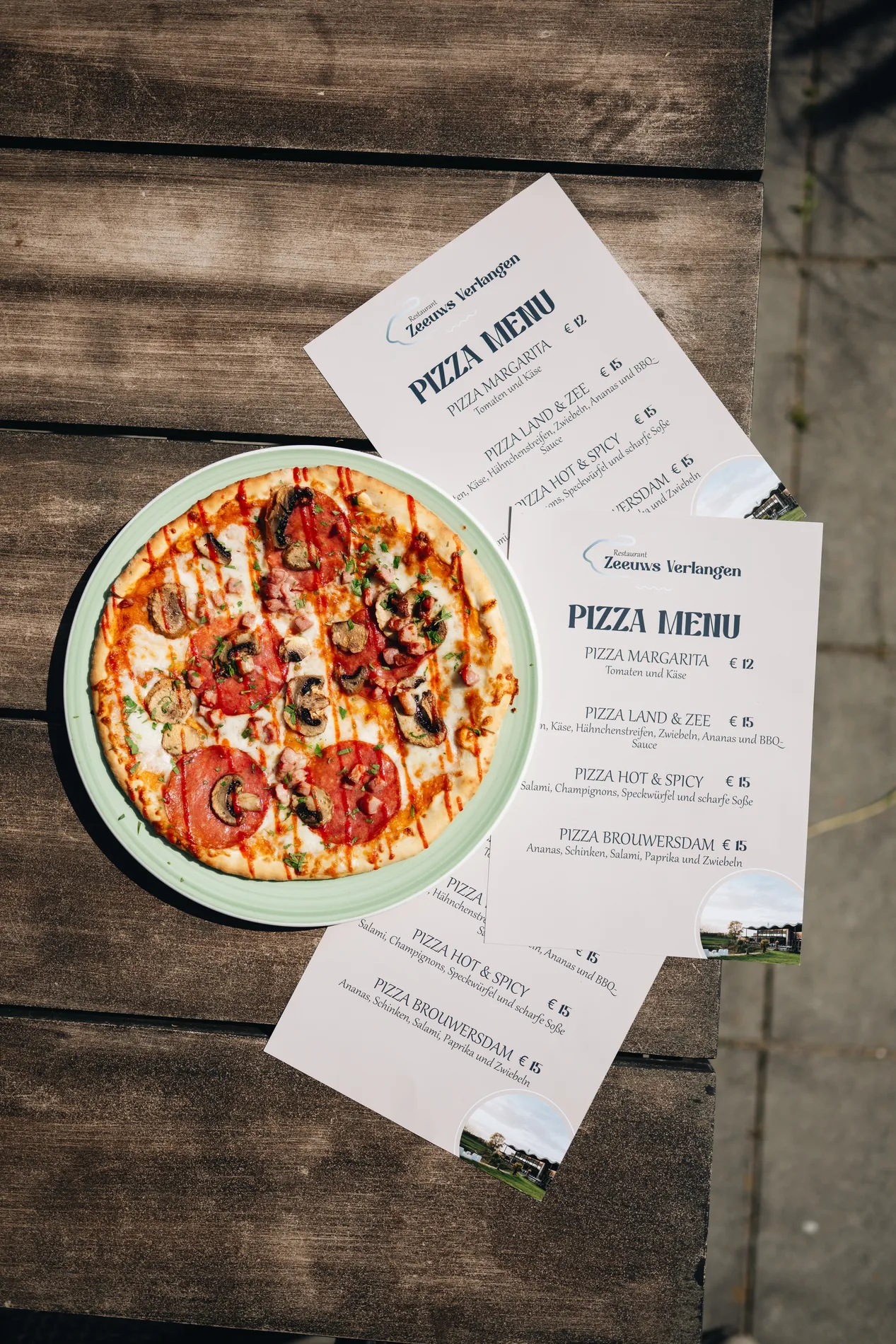 Een versgebakken, rijkelijk belegde pizza met salami en champignons op een lichtgroen bord, gepresenteerd naast de pizzamenukaarten van Restaurant Zeeuws Verlangen op een houten tafel.